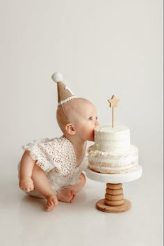 a baby with a party hat sniffing at a cake on a stand, while wearing a white dress