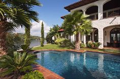 an outdoor swimming pool surrounded by palm trees and shrubs next to a large white house