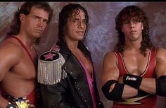 three men standing next to each other with their arms crossed and one man wearing a red wrestling suit