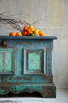 a green cabinet with fruit on top and branches in the background, against a gray wall