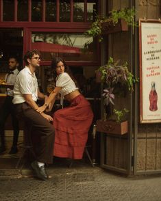 a man and woman sitting on a chair in front of a storefront with people standing around