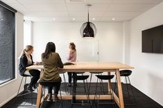 three women are sitting at a table in an office setting with a television on the wall