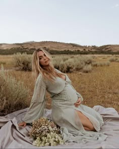 a pregnant woman sitting on top of a blanket in the middle of an open field