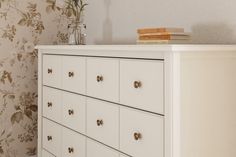 a white dresser sitting next to a wall with flowers on the top and bottom drawers