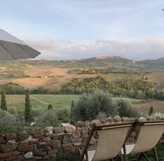 two chairs and an umbrella are sitting on the grass near a stone wall with rolling hills in the background