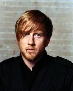 a man with red hair and blue eyes looks at the camera while standing in front of a brick wall