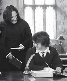 two people standing in front of a desk with a book on it and one person holding a pen