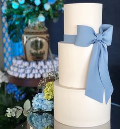 a three tiered white cake with blue ribbon on top and flowers in the background