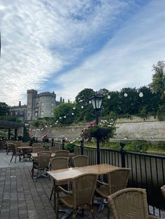 tables and chairs are lined up on the side walk near a river with buildings in the background