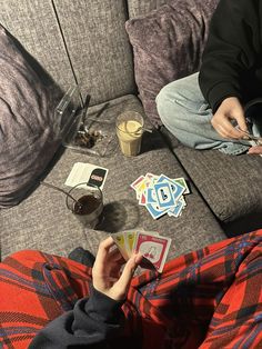 two people sitting on a couch playing cards with each other and drinking coffee in the background