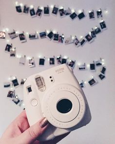 a person holding up a white camera in front of many polaroid pictures on the wall