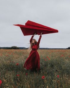 a woman in a red dress holds up a paper airplane above her head while standing in a field of poppies