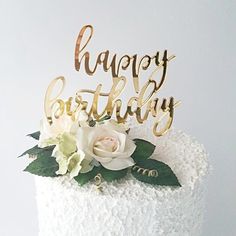 a white cake with gold lettering and flowers on the top that says, happy birthday