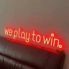 a neon sign that says we play to win on the wall above a leather couch