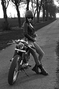 a woman sitting on top of a motorcycle wearing a helmet and leather jacket with trees in the background