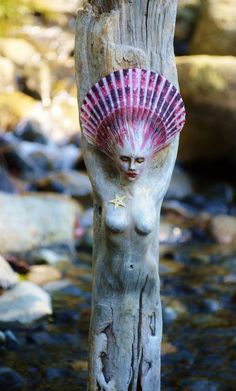 a wooden statue with a pink shell on it's head next to rocks and water