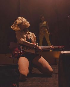 a woman sitting on the ground with a guitar in her hand and another person standing behind her