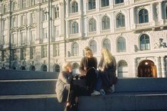 three women sitting on the steps in front of a large white building with arched windows