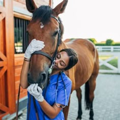 a woman in scrubs is petting a brown horse's head while standing next to a stable