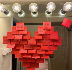 a bunch of red sticky notes hanging from a mirror with light bulbs above them that have been pinned to the ceiling