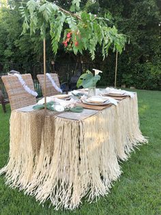 a table set up in the grass with plates and napkins on it, surrounded by greenery