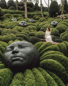 a woman sitting on top of a lush green field next to trees and bushes in the shape of heads