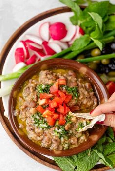 a person dipping some kind of food into a bowl with vegetables and herbs around it