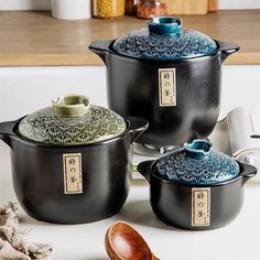 three black pots and two wooden spoons sitting on a counter top next to each other