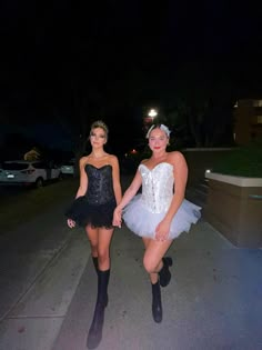 two women in short dresses are posing on the sidewalk at night with one holding the other's hand