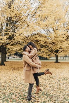 a man holding a woman in his arms while standing under trees with yellow leaves on the ground