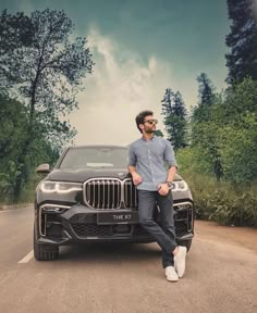 a man sitting on the hood of a bmw x7 parked in front of some trees