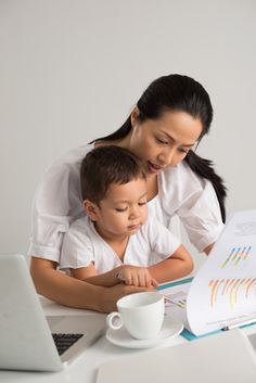 a woman and child are looking at a book on a table with a laptop in front of them