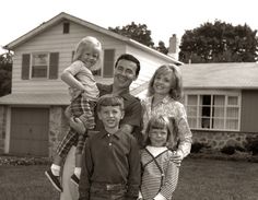 a black and white photo of a family standing in front of their house with the child on his shoulders