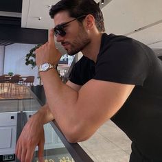 a man in sunglasses leaning on a railing looking at his watch while holding his hand to his face