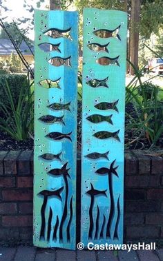 two metal fish sculptures sitting next to each other on a brick wall in front of a garden