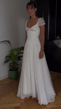 a woman in a white dress standing on a wooden floor next to a potted plant