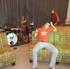 a man sitting on top of a couch in front of a drum set and another person standing behind him