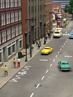 toy cars are driving down the street in front of tall buildings and people walking on the sidewalk