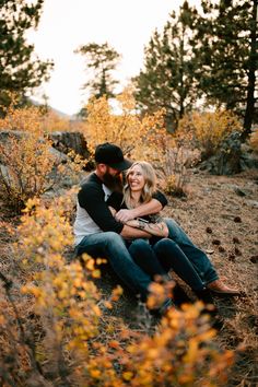 a man and woman are sitting on the ground in front of some trees with their arms around each other