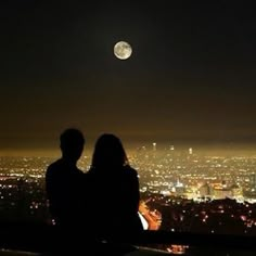 two people are sitting on a ledge looking at the city lights in the night sky