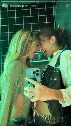 two women standing next to each other in front of a green tiled wall and one holding a cell phone