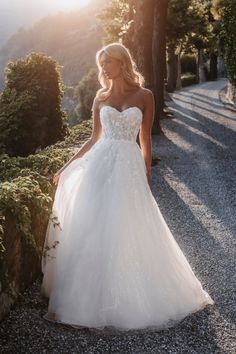 a woman in a white wedding dress standing on gravel road with trees and bushes behind her