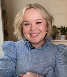 a woman with blonde hair wearing a blue shirt and smiling at the camera while sitting on a couch