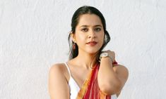 a woman wearing a red and orange sari with her hands on her hips, leaning against a white wall