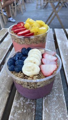 two plastic bowls filled with fruit and granola on top of a wooden picnic table