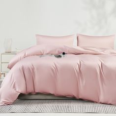 a bed with pink sheets and pillows in a white room next to a nightstand table