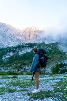 a woman standing on top of a lush green hillside next to a mountain covered in clouds