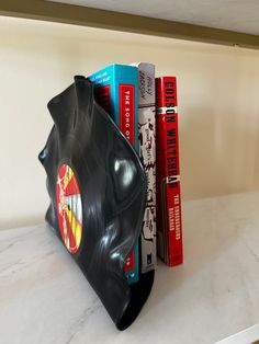 there is a black plastic bag on top of the bookshelf with red and blue books