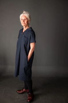 an older woman standing in front of a dark background wearing a blue dress and brown shoes