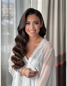 a woman with long brown hair wearing a white dress and smiling at the camera while standing in front of a window